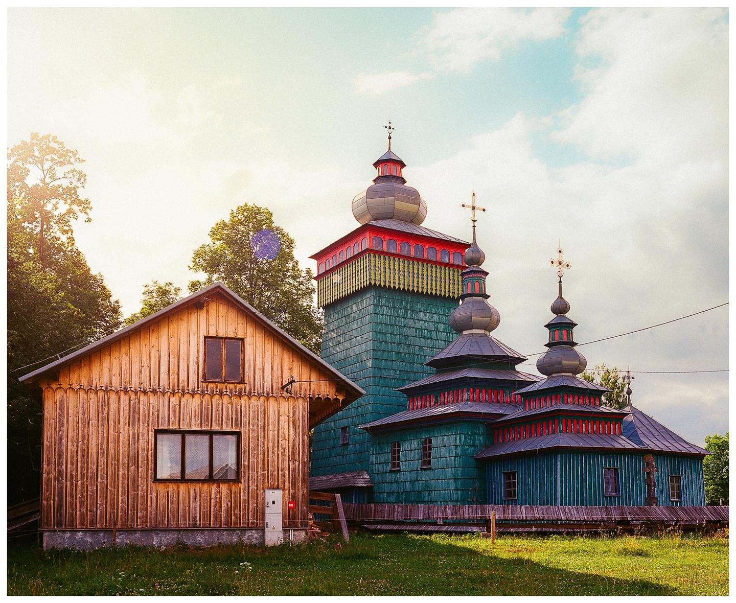A wooden church with multiple domes and a cross on each dome stands next to a wooden house in a grassy field.