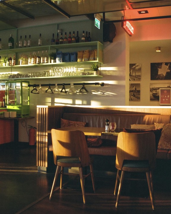 Sunlight illuminates the interior of a bar with shelves of bottles and glasses, and a seating area with chairs and a booth.