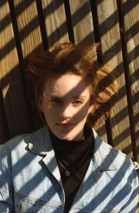 A woman with short hair is lying on a wooden surface, looking up at the camera, with shadows from a slatted structure falling across her face and body.