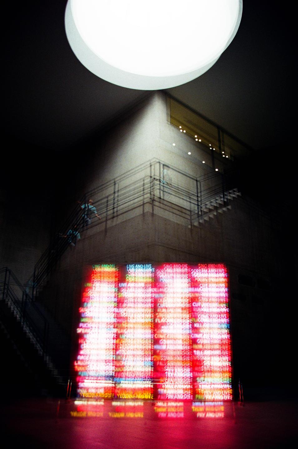 A large, bright circular light illuminates a dark interior space with a glowing red and yellow neon sign at the bottom.