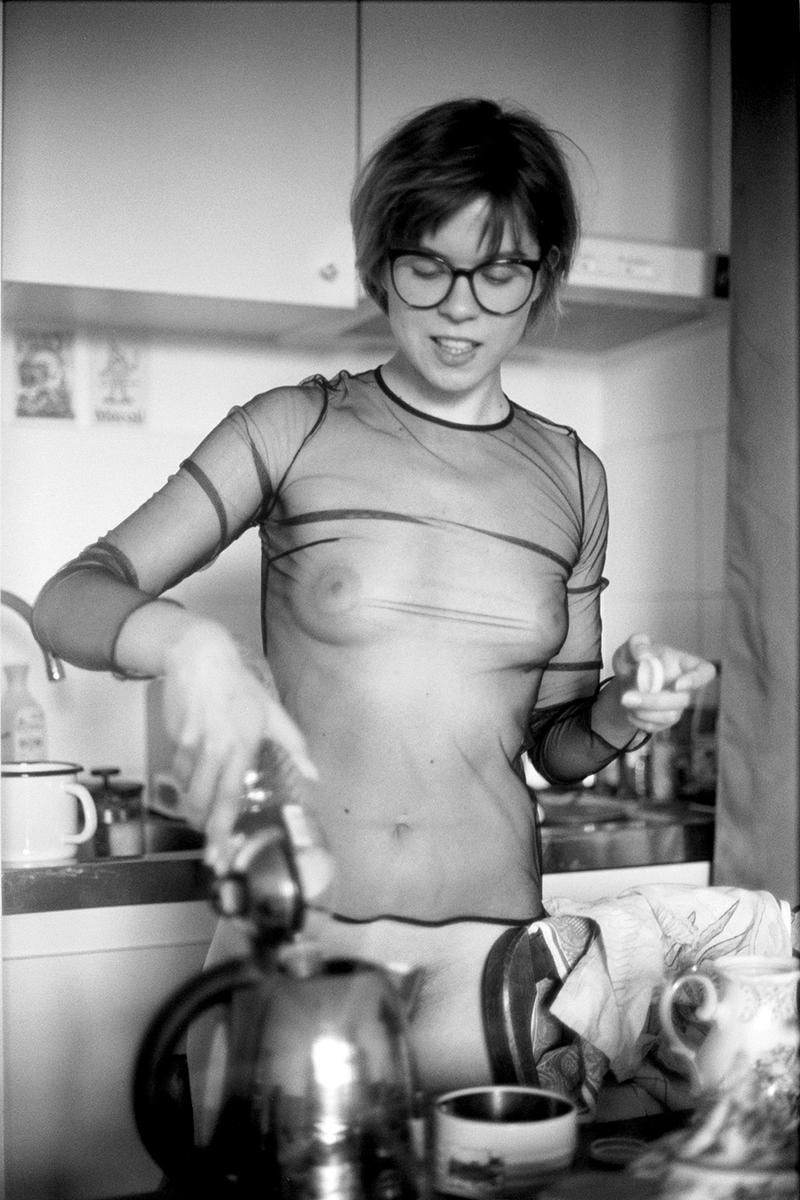 A woman in a sheer top and glasses is in a kitchen, holding a small white object in one hand and operating a coffee maker with the other.