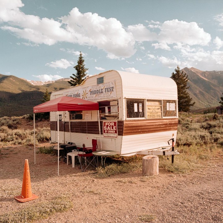 A white and brown camper trailer with a red canopy is parked in a dry, grassy field with mountains in the background.