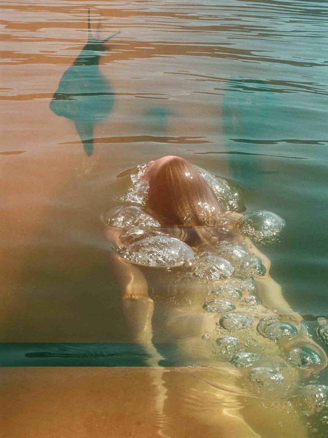 A person's head and shoulder are visible above and below the surface of the water, with bubbles around them.