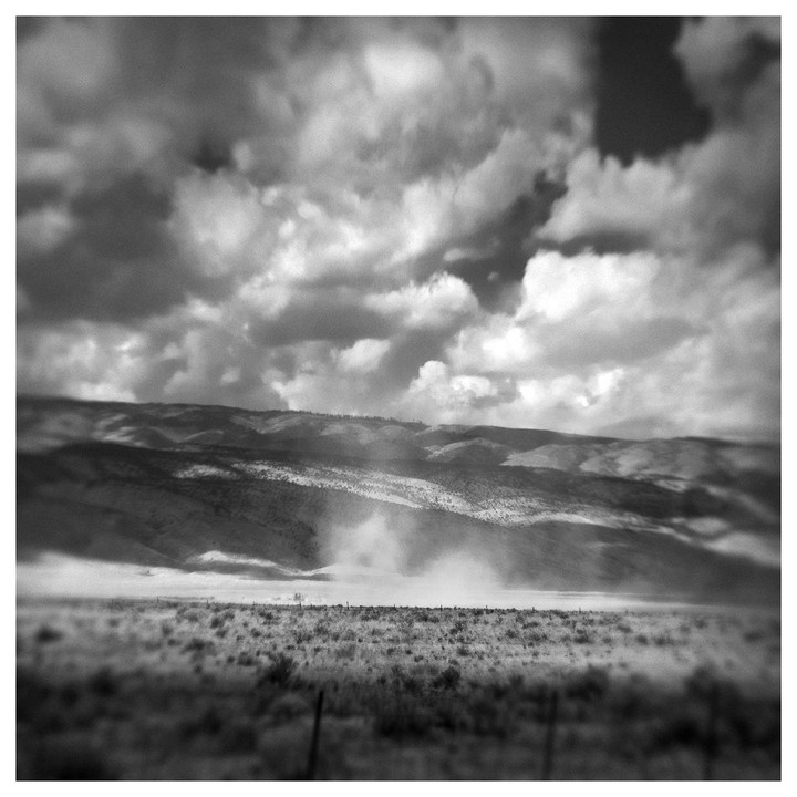 A dust devil swirls in a vast, dry field with mountains in the background under a cloudy sky.