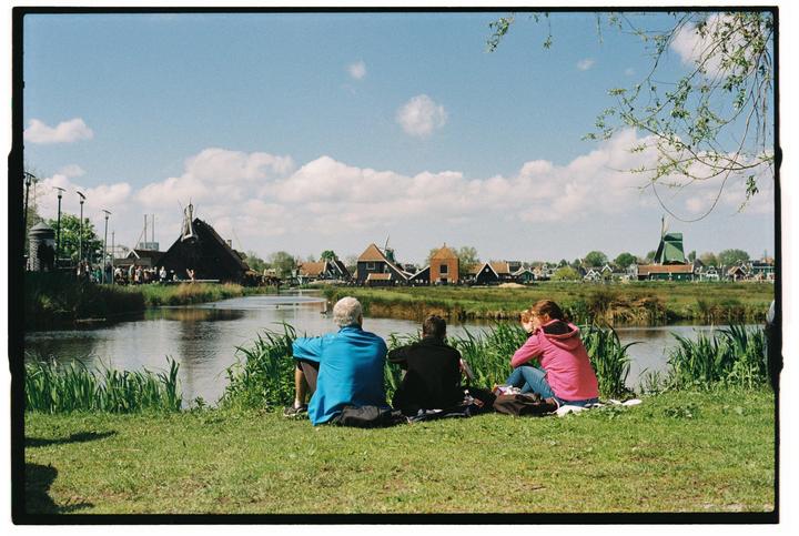 Three people sit on a grassy bank overlooking a canal with traditional Dutch buildings and a windmill in the background.