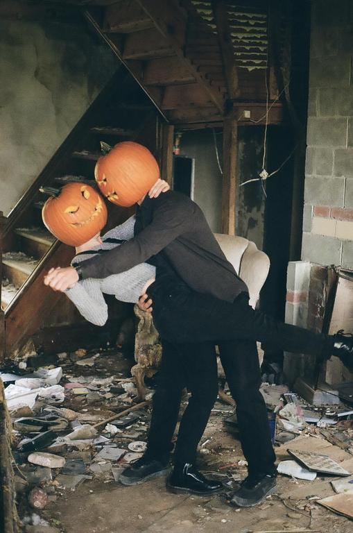 A couple with carved pumpkins on their heads dances in a dilapidated room with debris on the floor.