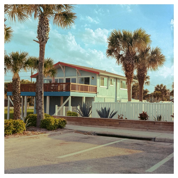 A light blue house with a red roof and a wooden balcony is surrounded by palm trees and a white fence.