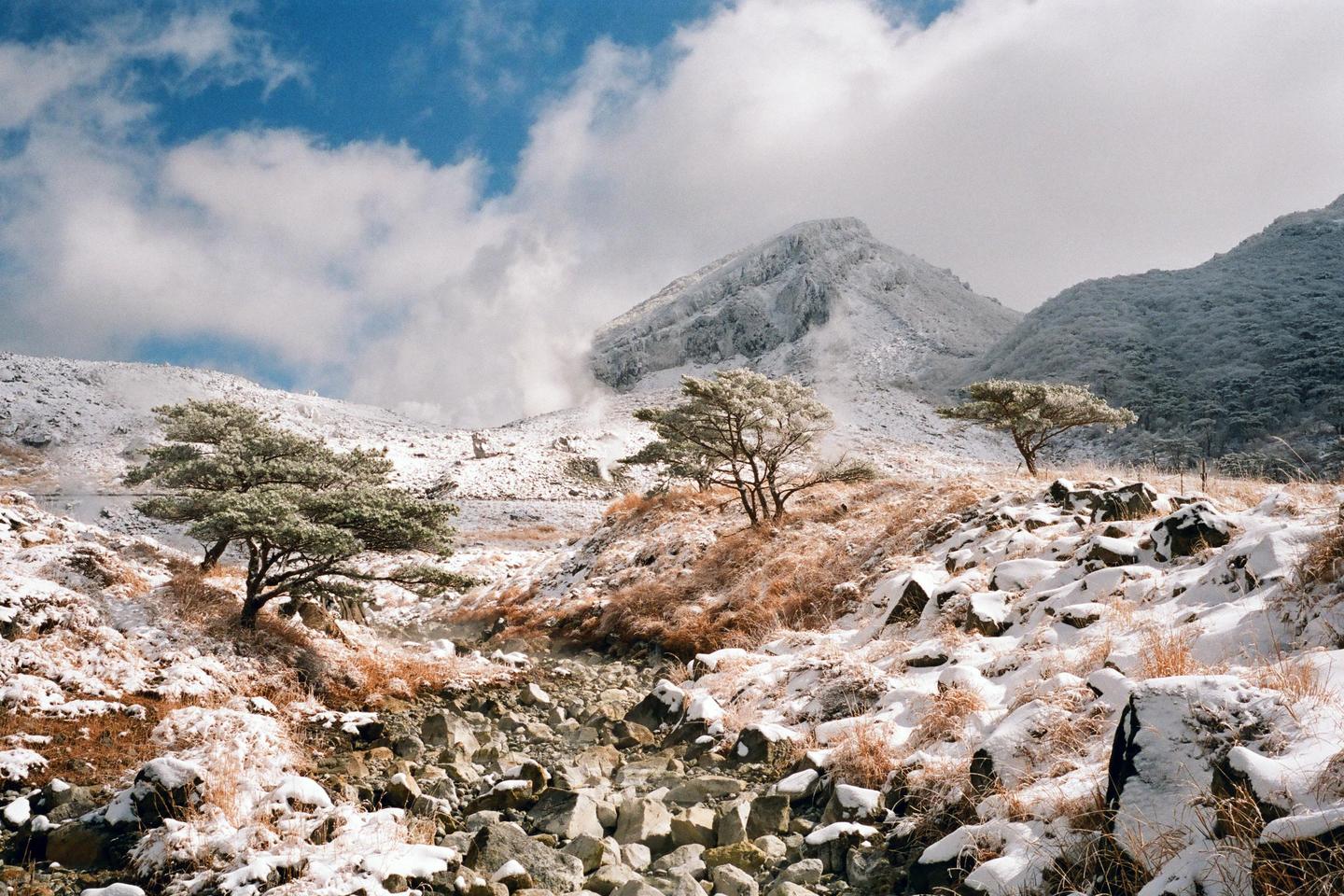 A snow-covered mountain landscape features a rocky stream bed, scattered trees, and a prominent peak under a cloudy sky.