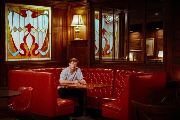 A man sits alone at a table in a restaurant booth with red upholstery and stained glass windows.