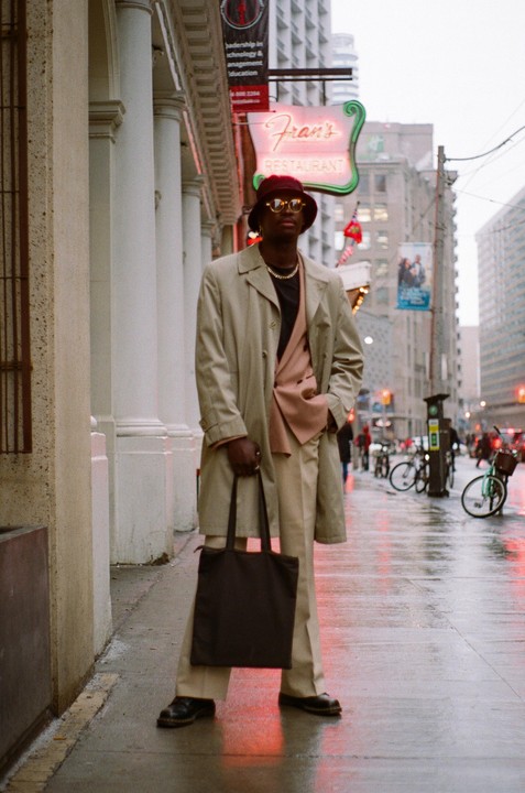 A man in a trench coat and bucket hat stands on a wet city street next to a restaurant sign.