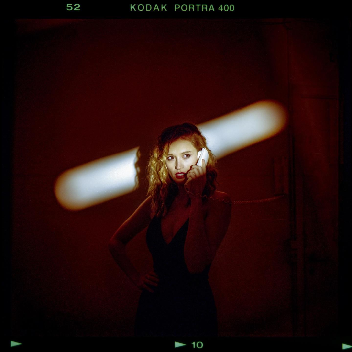 A woman in a dark dress holds a telephone receiver to her ear in a dimly lit room with a bright light streak on the wall.