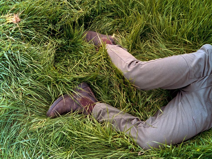 A person wearing brown boots and gray pants is lying in tall green grass.