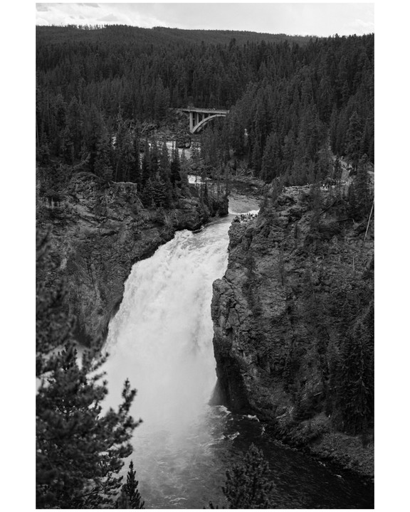 A large waterfall cascades into a river, surrounded by a dense forest and a bridge in the distance.