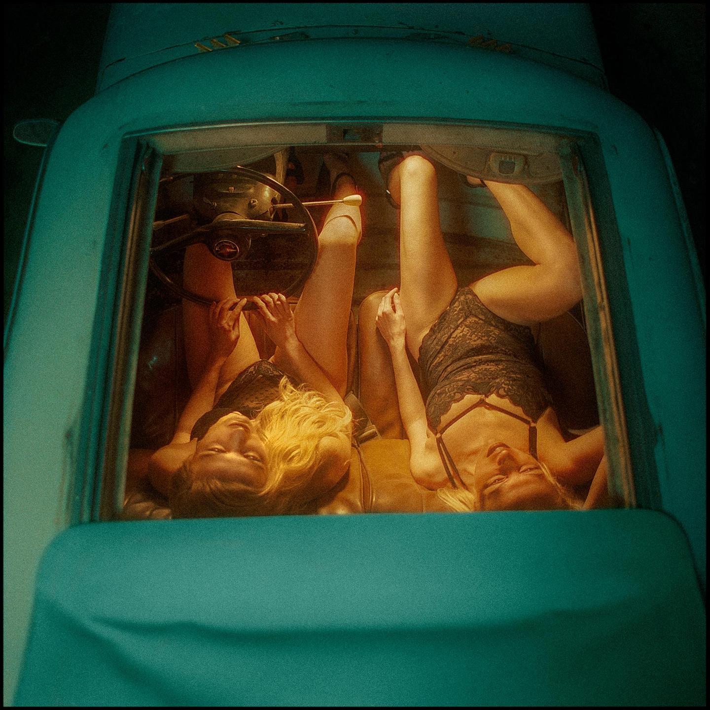 Two women in lingerie are lying in the back of a teal car.