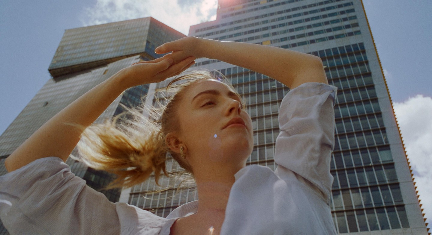 A woman with her arms raised above her head looks up at the sky with a tall building in the background.