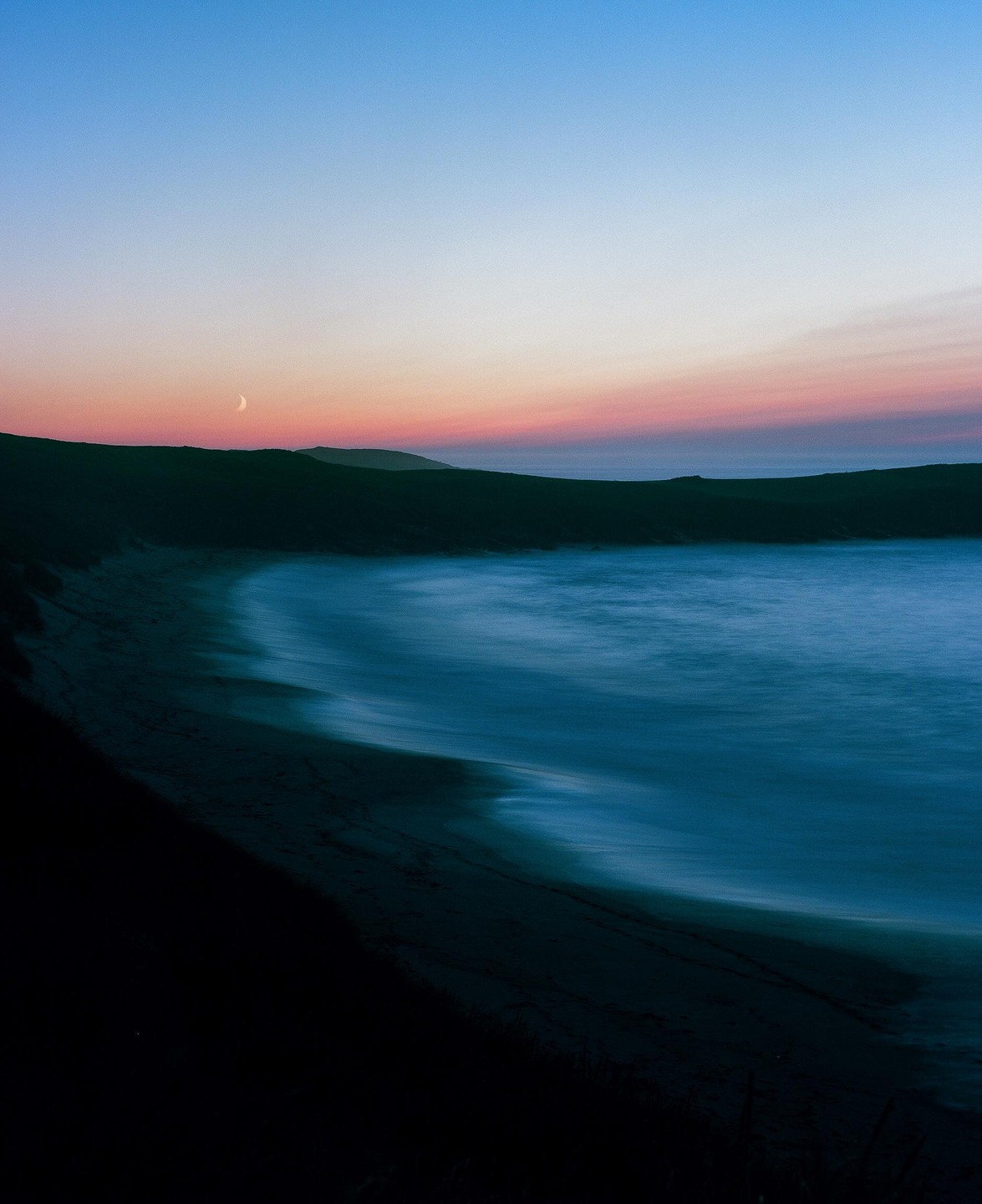 A crescent moon hangs over a bay with a sandy beach and hills under a colorful sky at dusk.