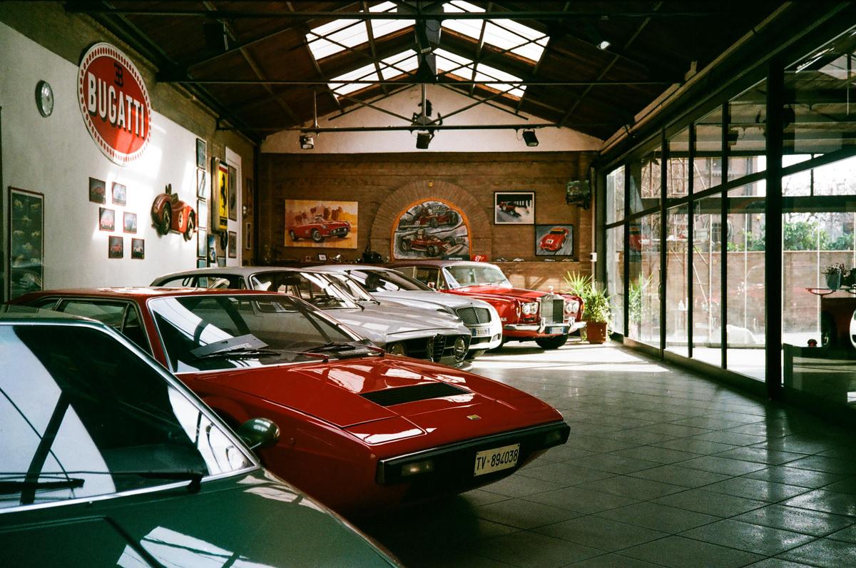A showroom with several vintage cars, including a red sports car in the foreground, is illuminated by natural light from skylights and large windows.