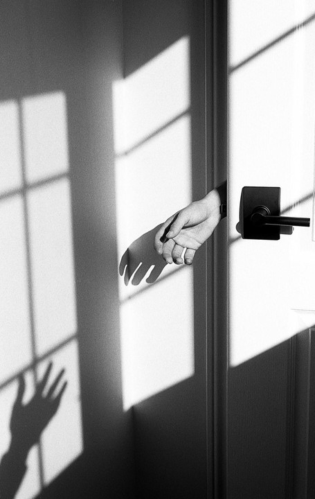 A hand reaches out from behind a door, casting a shadow on the wall with window light.