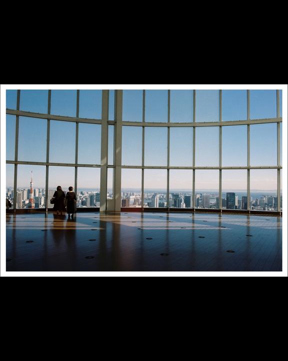 Two people stand in front of a large window overlooking a city skyline with a prominent tower.