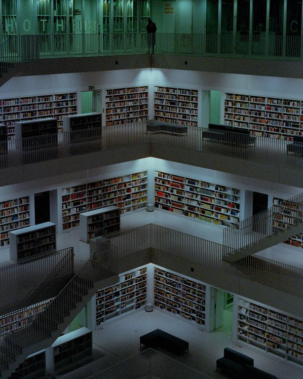 A person stands on an upper level overlooking a multi-story library with many bookshelves and staircases.