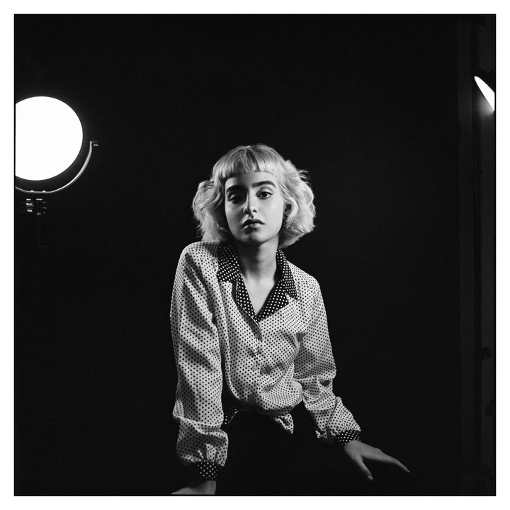 A woman with short, curly hair and a polka dot shirt sits in a studio with two lights.