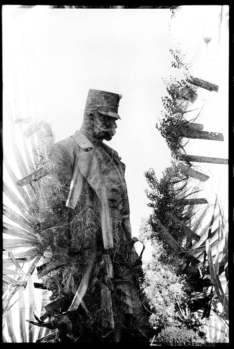 A double exposure shows a statue of a man in uniform overlaid with foliage and wooden planks.