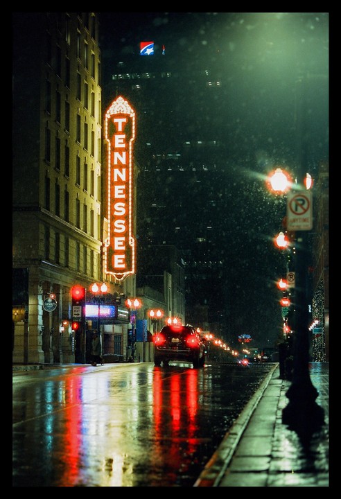 A neon sign for "Tennessee" illuminates a wet street at night, reflecting in the puddles.