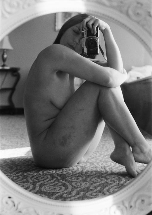 A woman sits nude on a patterned carpet, reflected in an ornate mirror, holding a camera to her face.