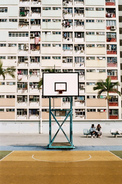 A basketball hoop stands on a court in front of a large apartment building with two people sitting on a bench.