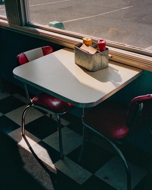 A diner booth with a table and two chairs sits next to a window, with a condiment caddy on the table.