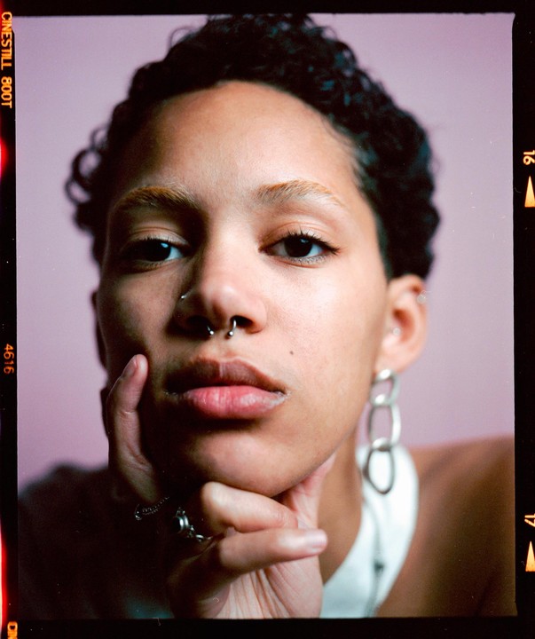A woman with short curly hair and bleached eyebrows rests her chin on her hand, looking directly at the viewer.