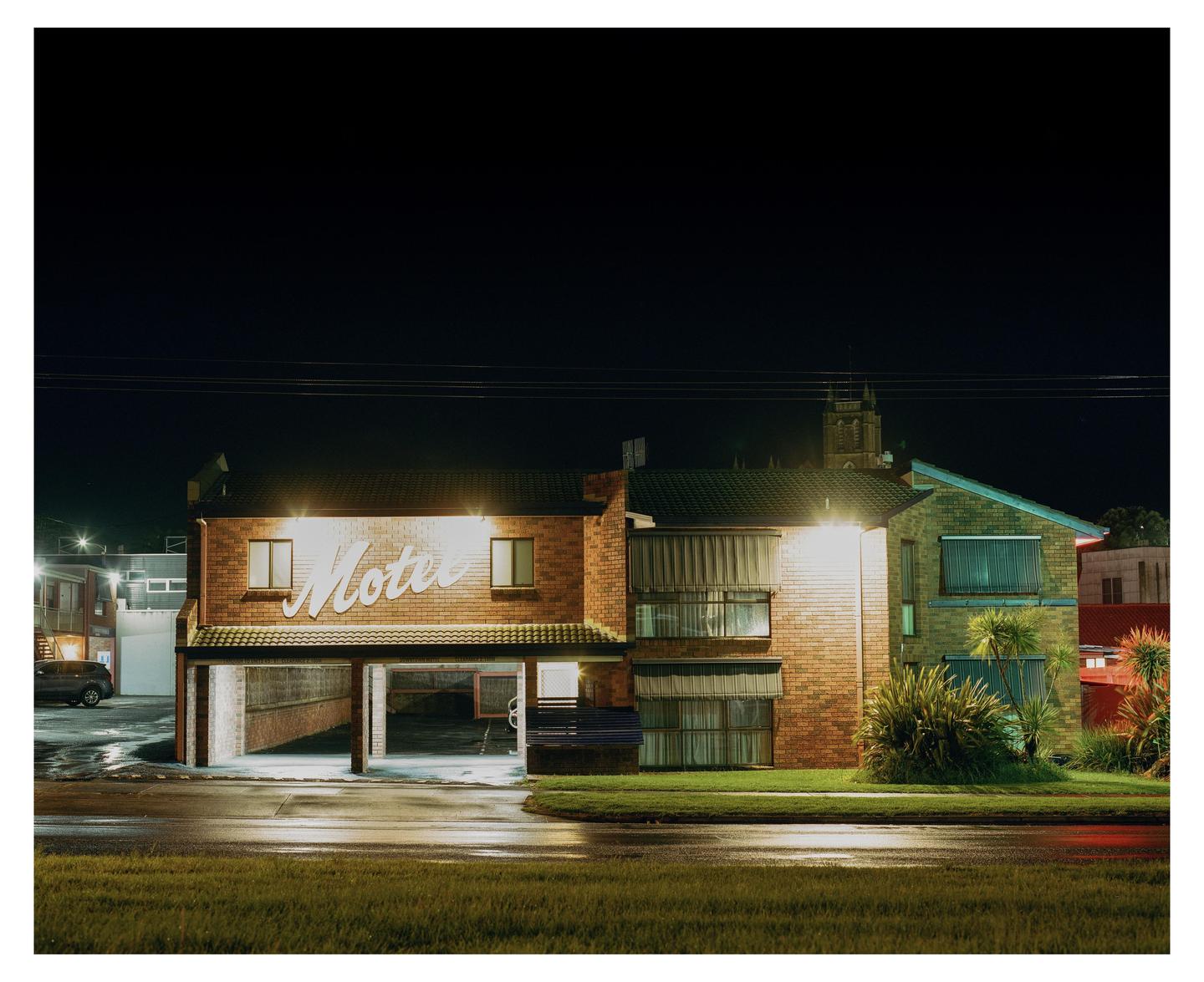 A motel with an illuminated sign is visible at night, with a wet street in the foreground.