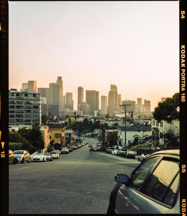 A city skyline is visible in the distance over a residential street lined with parked cars.
