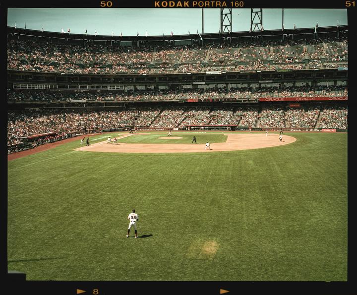 A baseball player stands on a green field with a stadium full of spectators in the background.