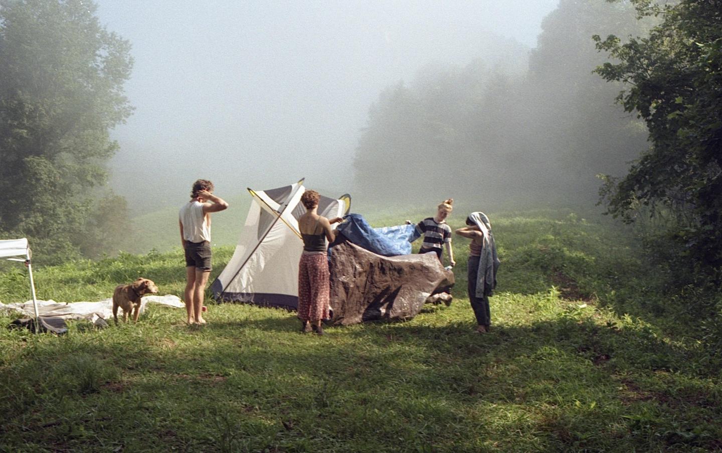 Four people and a dog are gathered around a tent in a grassy, hazy field.
