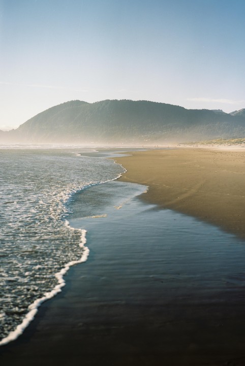 Waves crash on a sandy beach with a large, forested mountain in the background.