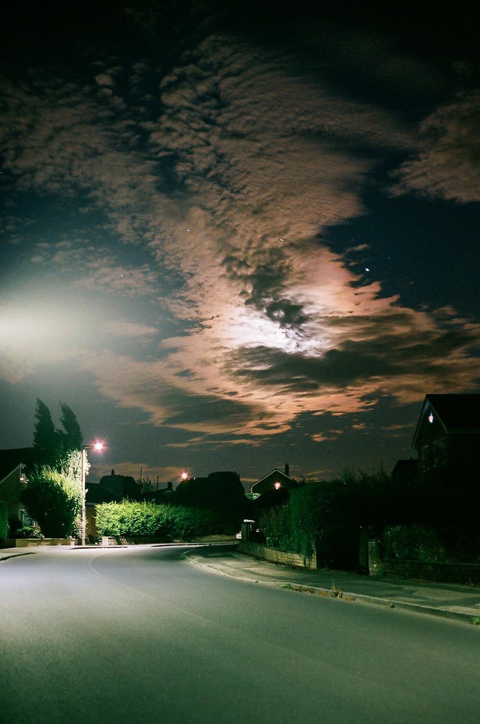 A full moon illuminates the clouds above a residential street at night.