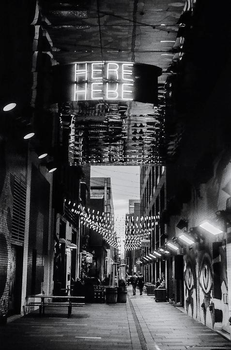 A neon sign reading "HERE HEBE" hangs above a narrow city street decorated with string lights and graffiti.