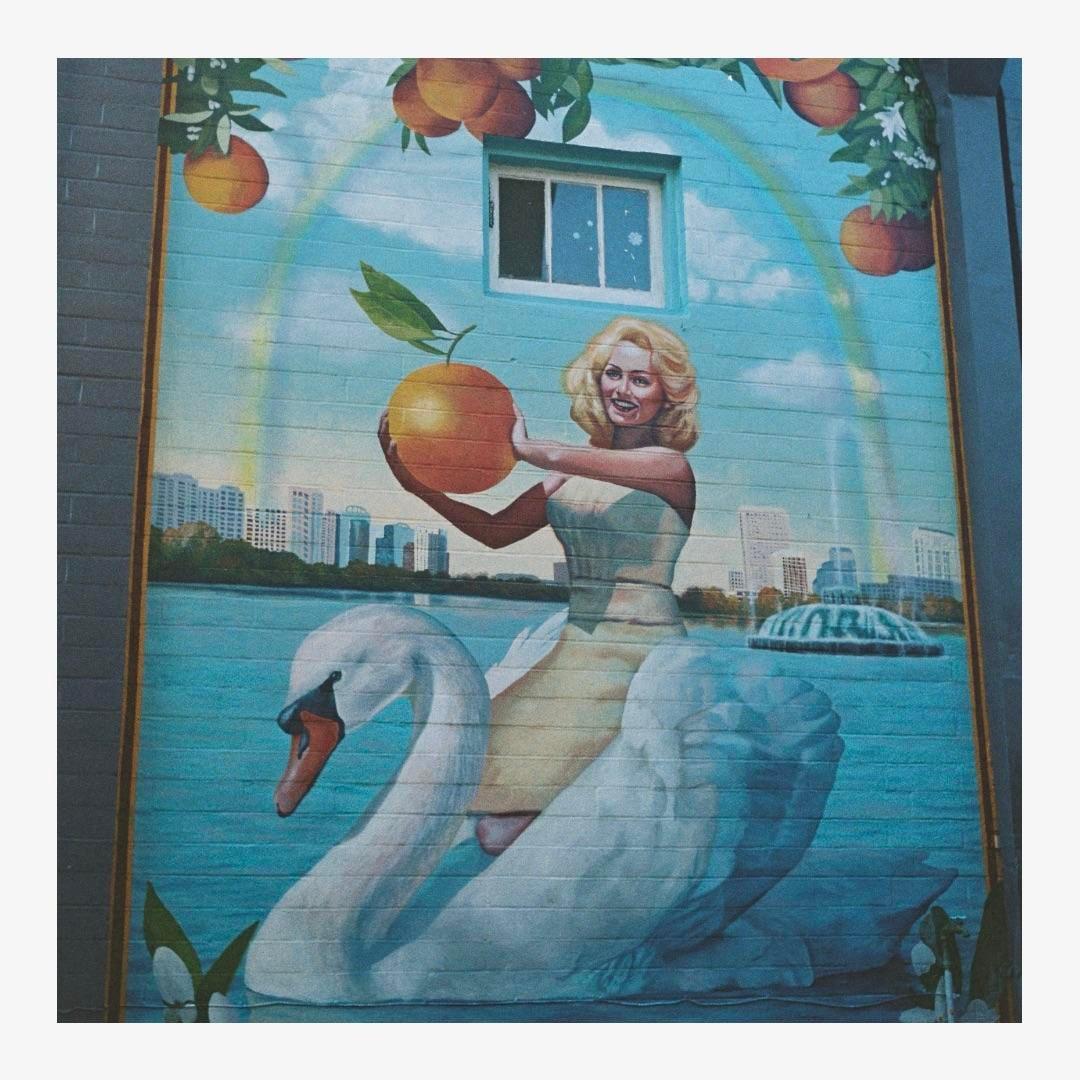 A mural depicts a woman riding a swan on water, holding an orange, with a city skyline and oranges in the background.