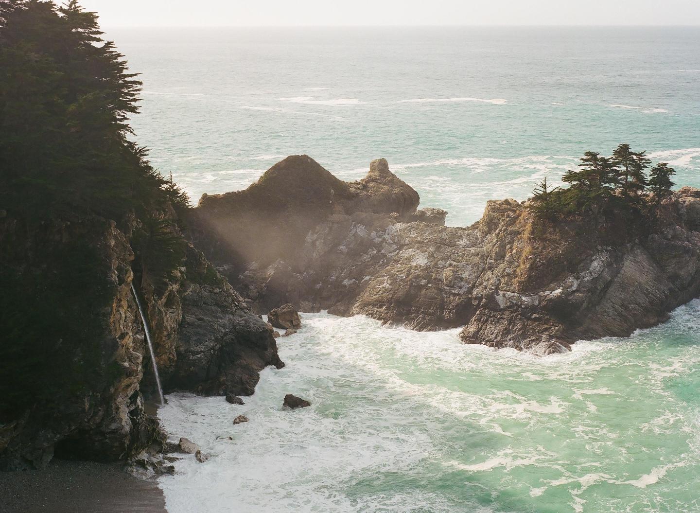 A waterfall cascades from a tree-covered cliff into the ocean, with waves crashing against rocky formations.
