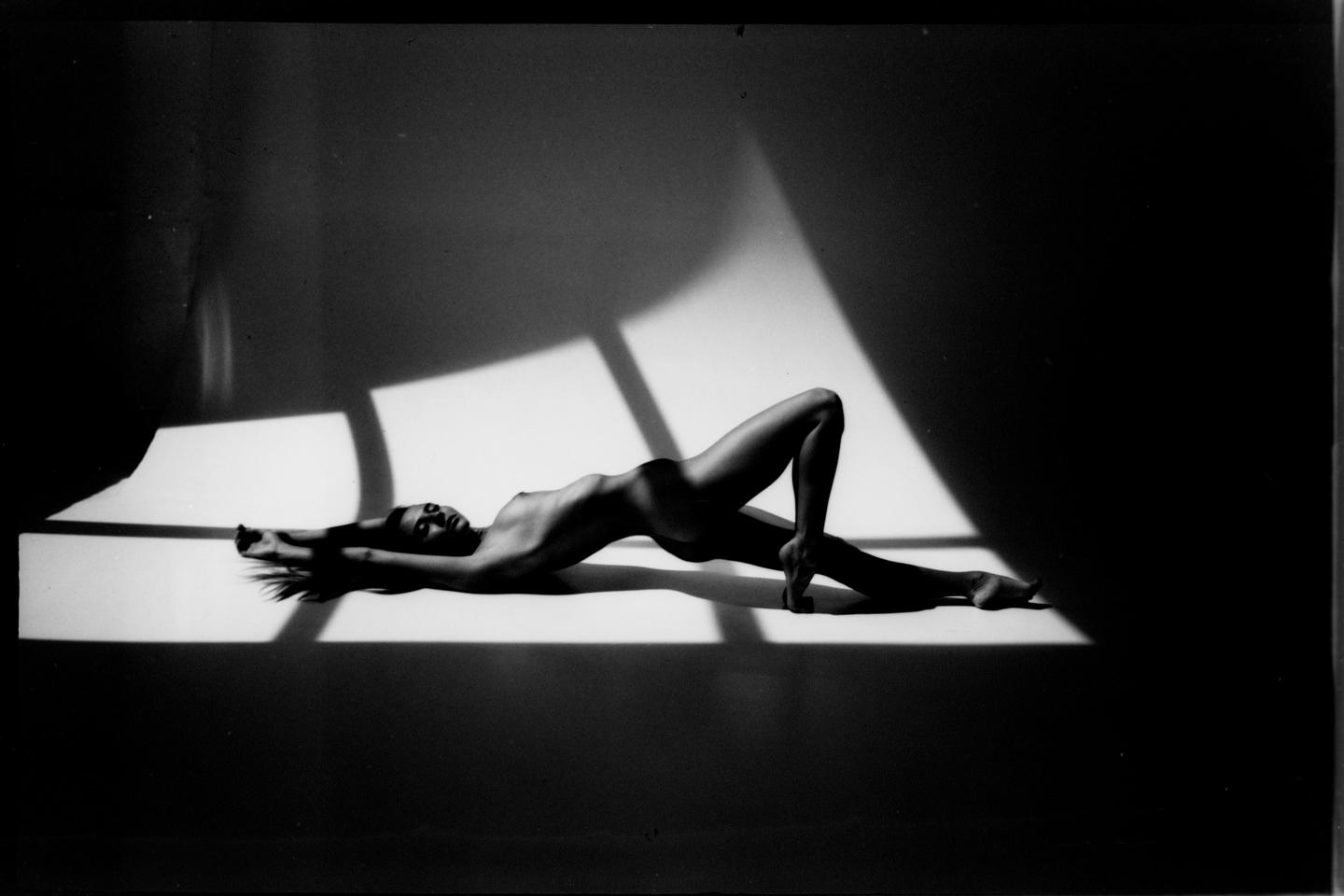 A nude woman lies on her back with her arms stretched above her head and her legs bent, with light and shadow creating geometric shapes around her.
