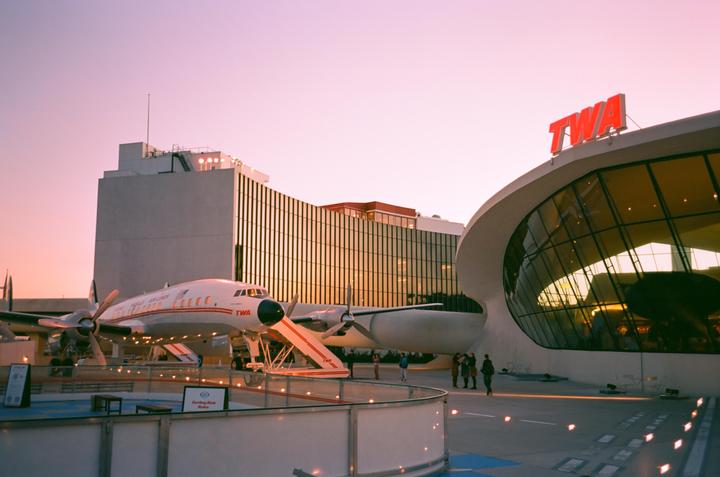 A TWA airplane is parked next to a modern building with a large glass facade under a pink sky.