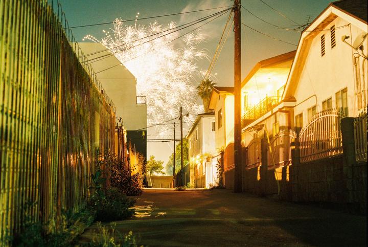 Fireworks explode over houses and a fence in an alley at night.