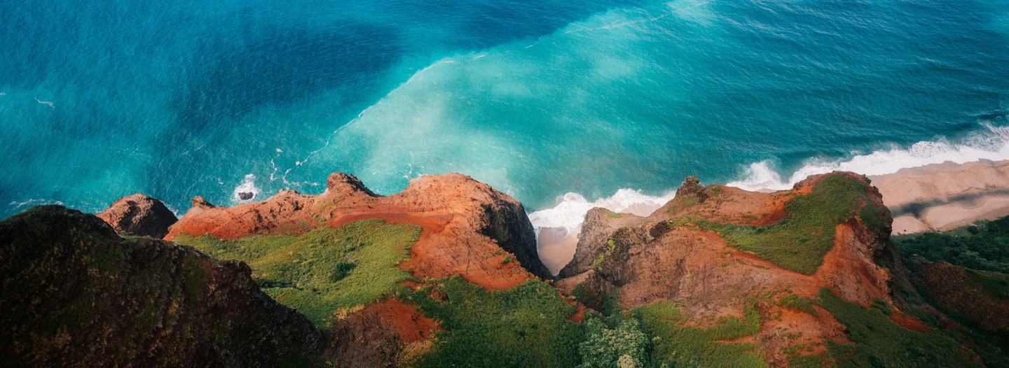 An aerial view shows a rugged coastline with red cliffs and green vegetation next to a turquoise ocean with waves crashing on a sandy beach.