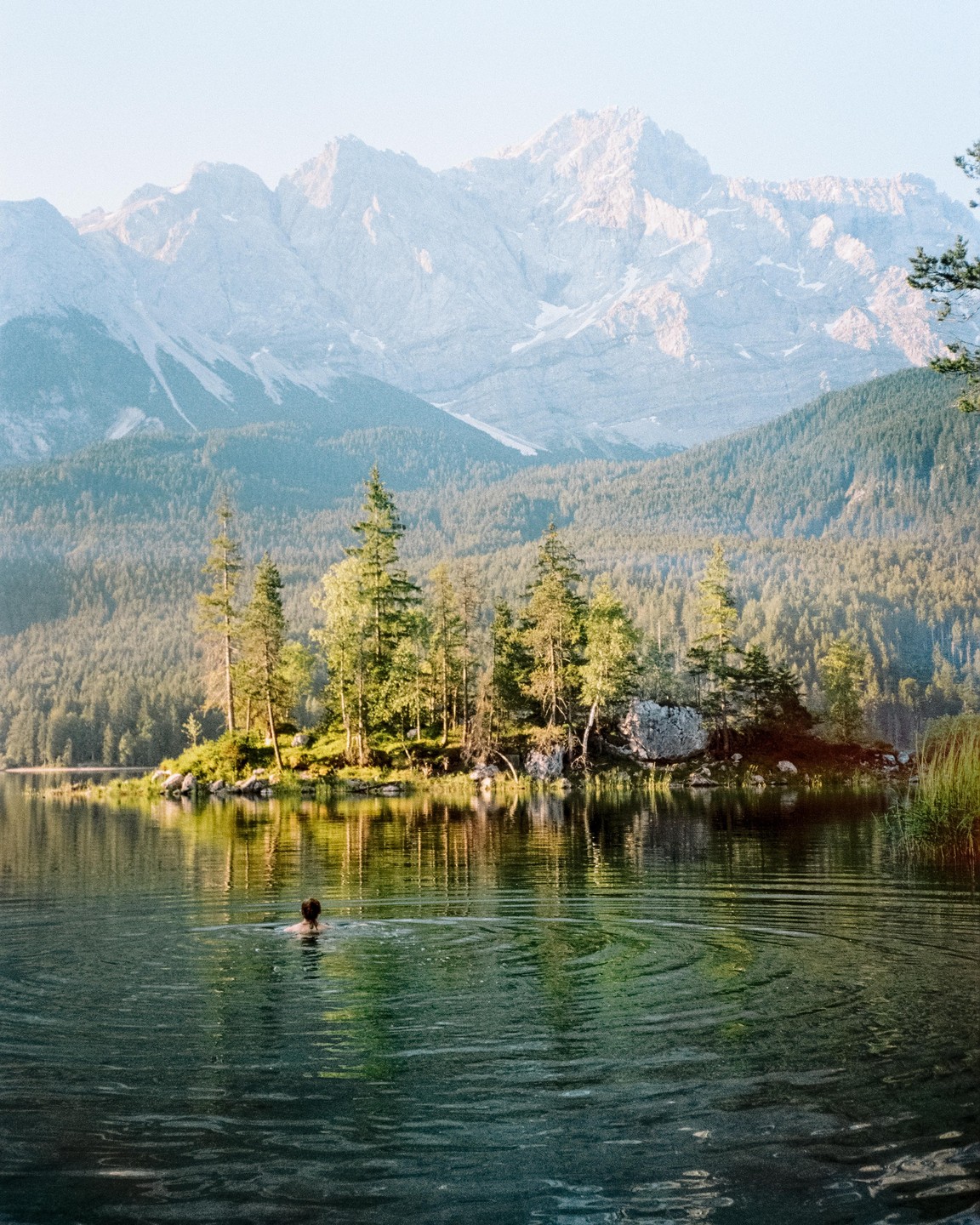 A person swims in a lake with a small island of trees and mountains in the background.