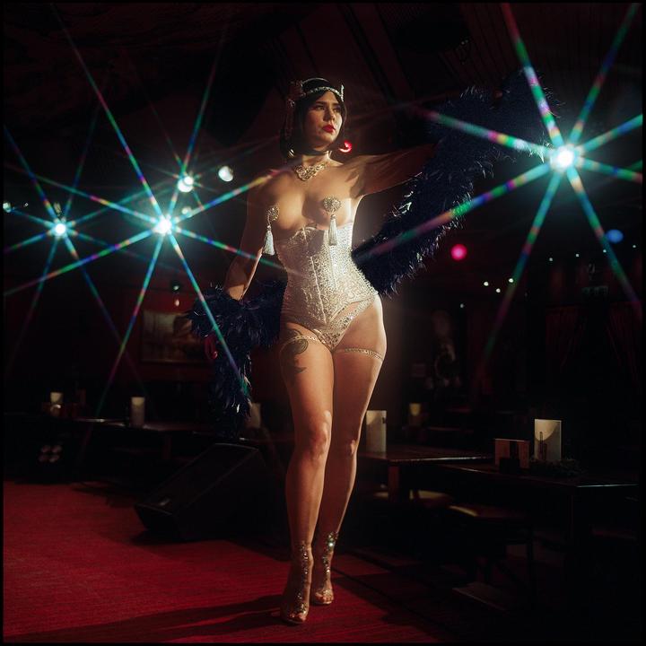 A woman in a white corset and feathered boa stands on a red carpet stage with bright lights in the background.
