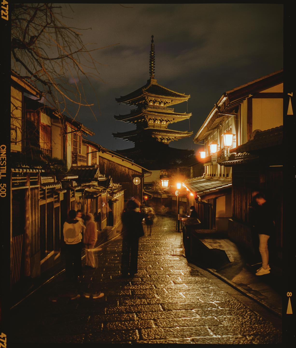 A pagoda rises above a narrow, wet street lined with traditional buildings and people at night.