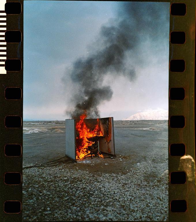 A burning refrigerator emits thick black smoke into the sky in a desolate, rocky landscape with a distant mountain.