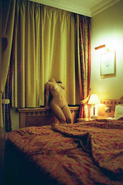 A nude woman leans against a window with yellow curtains in a hotel room with a patterned bedspread.