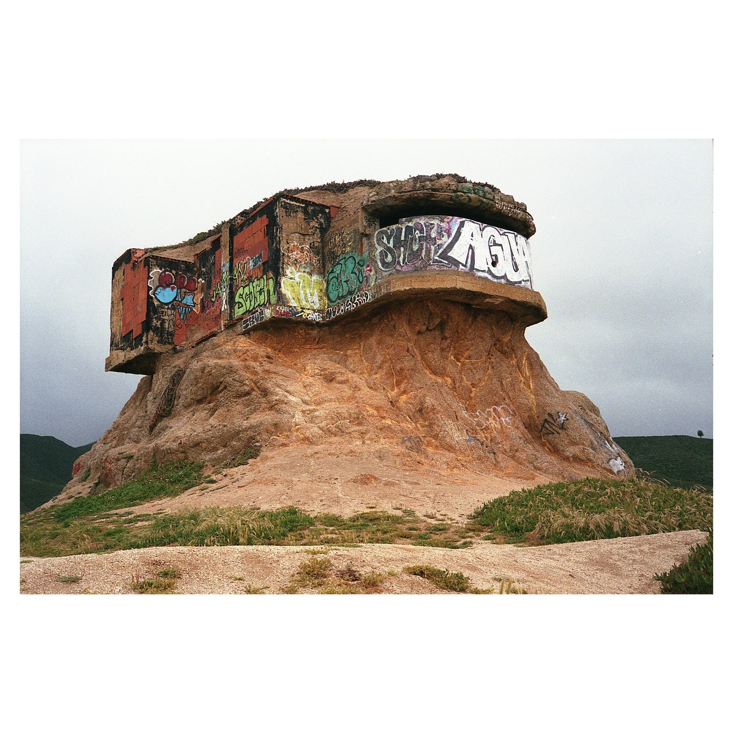 A concrete bunker covered in graffiti sits atop a rocky hill under a cloudy sky.